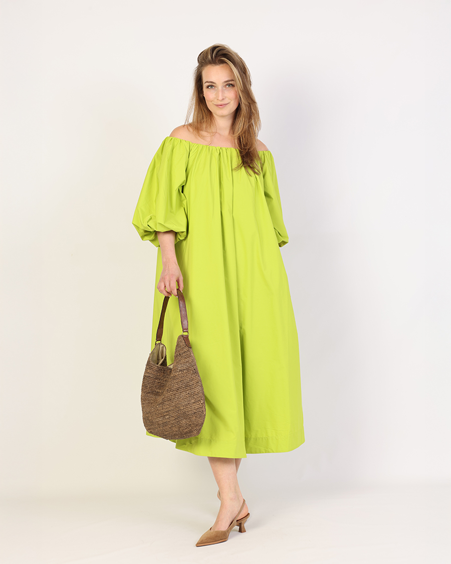 Essentiel Antwerp jurk: Zomerse Mode 2025 Vrouw draagt een opvallende limoengroene off-shoulder jurk met volumineuze halflange mouwen en een wijde pasvorm, gecombineerd met een naturel geweven handtas en elegante, spitse muiltjes met lage hak, perfect voor een zomerse, casual look.