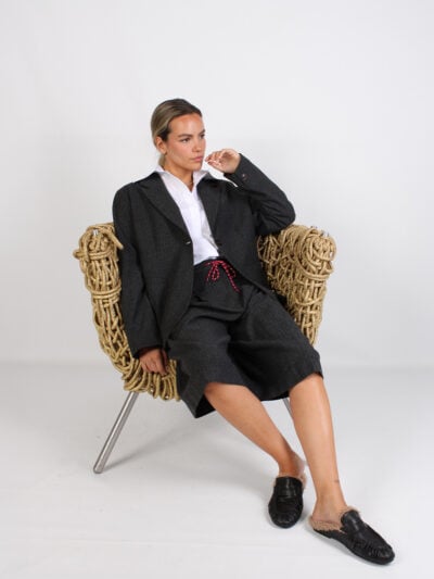 Zittend model in donkergrijs colbert, witte blouse, bermuda en zwarte instappers